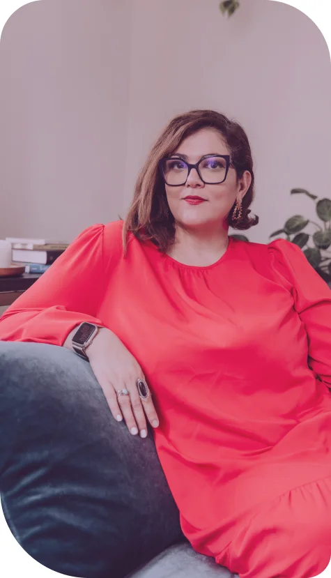 Picture of Mariel, a latin woman, with a round face, red lipstick and glasses, wearing a coral dress and posing in a couch